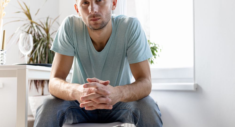 serious young man in t-shirt and jeans sits on a chair in a bright room