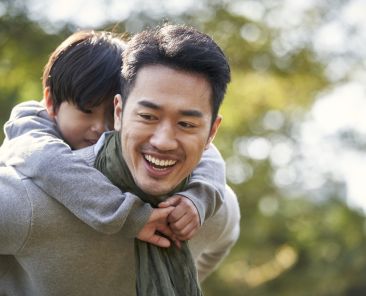 asian father carrying son on back having fun outdoors in park