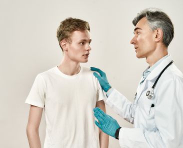 Meeting at hospital. Mature professional doctor in medical uniform and blue sterile gloves speaking with worried young male patient, explaining diagnosis. Healthcare, medicine. Medical consultation