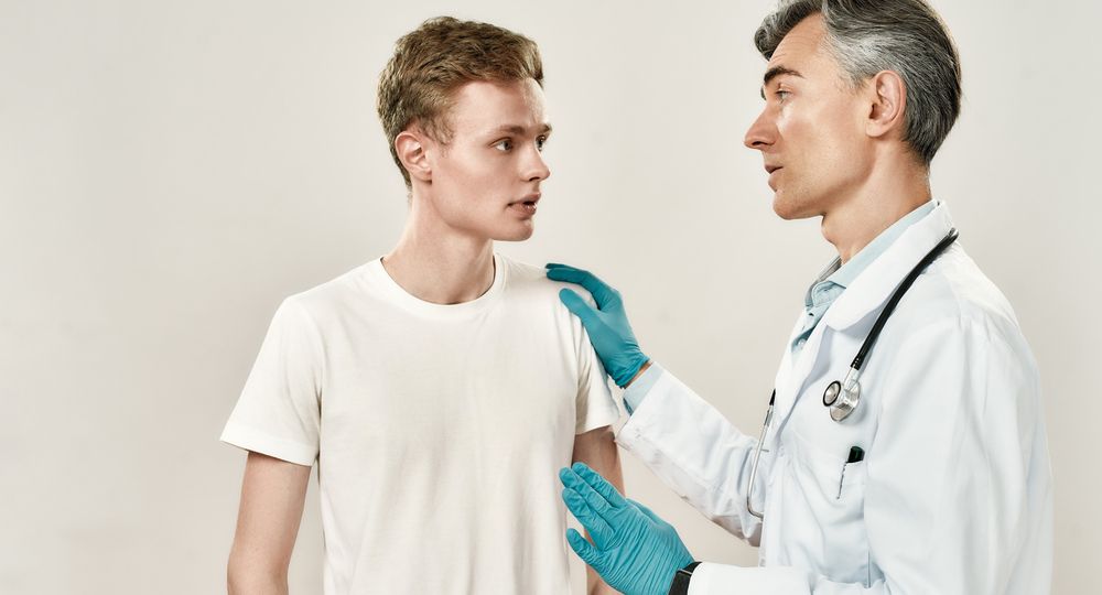 Meeting at hospital. Mature professional doctor in medical uniform and blue sterile gloves speaking with worried young male patient, explaining diagnosis