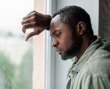 Unhappy frustrated adult african american guy in casual suffering from depression and bad news near window in home interior. Health problems, stress from self-isolation during covid-19 quarantine