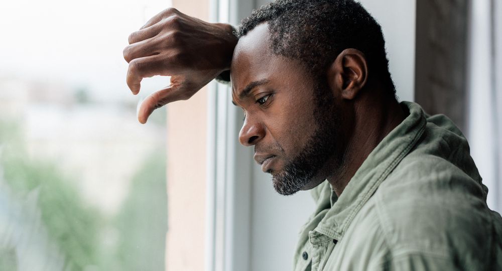 Unhappy frustrated adult african american guy in casual suffering from depression and bad news near window